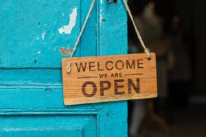A rustic wooden 'Welcome We Are Open' sign hanging on a vibrant blue door.